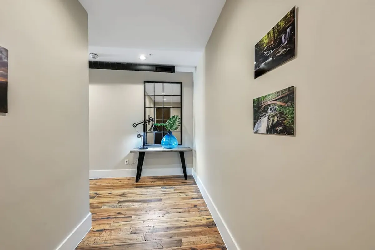 A serene workspace featuring wooden flooring, a desk with a lamp perfect for productivity, a calming blue glass vase, and wall-mounted photos that add a touch of character. The natural lighting and thoughtful decor create an inviting setting for work or creative pursuits.