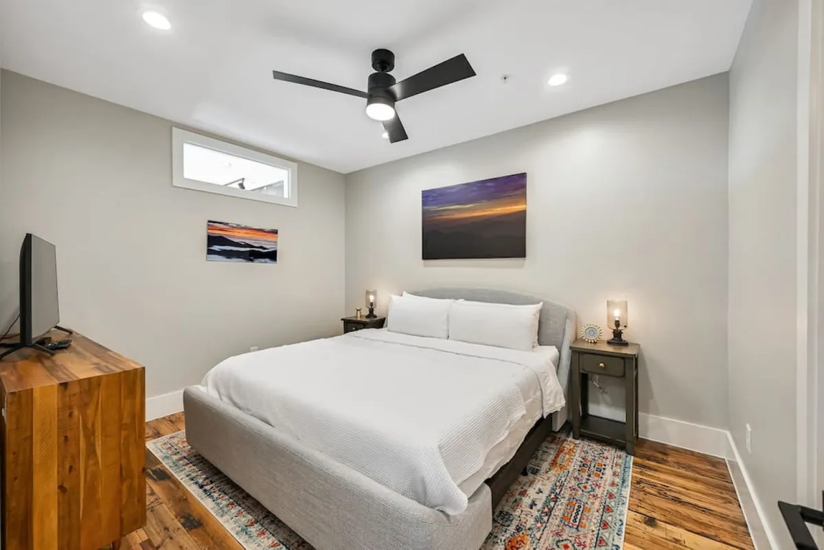 A tranquil bedroom scene with a king bed, soothing artwork, chic nightstands, a television, and a cozy rug underfoot. The ambiance is peaceful and inviting, perfect for winding down after a busy day. The layering of soft textures and thoughtful design details create a sense of warmth and comfort.
