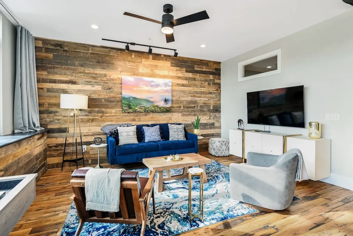 This image centers on a blue sofa set against a wood accent wall, complemented by colorful artwork and a contemporary area rug. The TV unit and soft lighting add to the relaxed, stylish atmosphere. Sunlight filtering through the side hints at large windows, and the overall decor bridges modern touches with nods to the building's historic character.