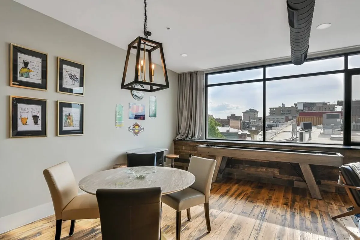 The dining area boasts a wooden table with a bench and chairs, positioned near a large window that bathes the room in natural light. Decorative artwork on the walls highlights local influences, while the clean lines of the furnishings underscore a contemporary aesthetic. The setting feels inviting for both meals and conversation.