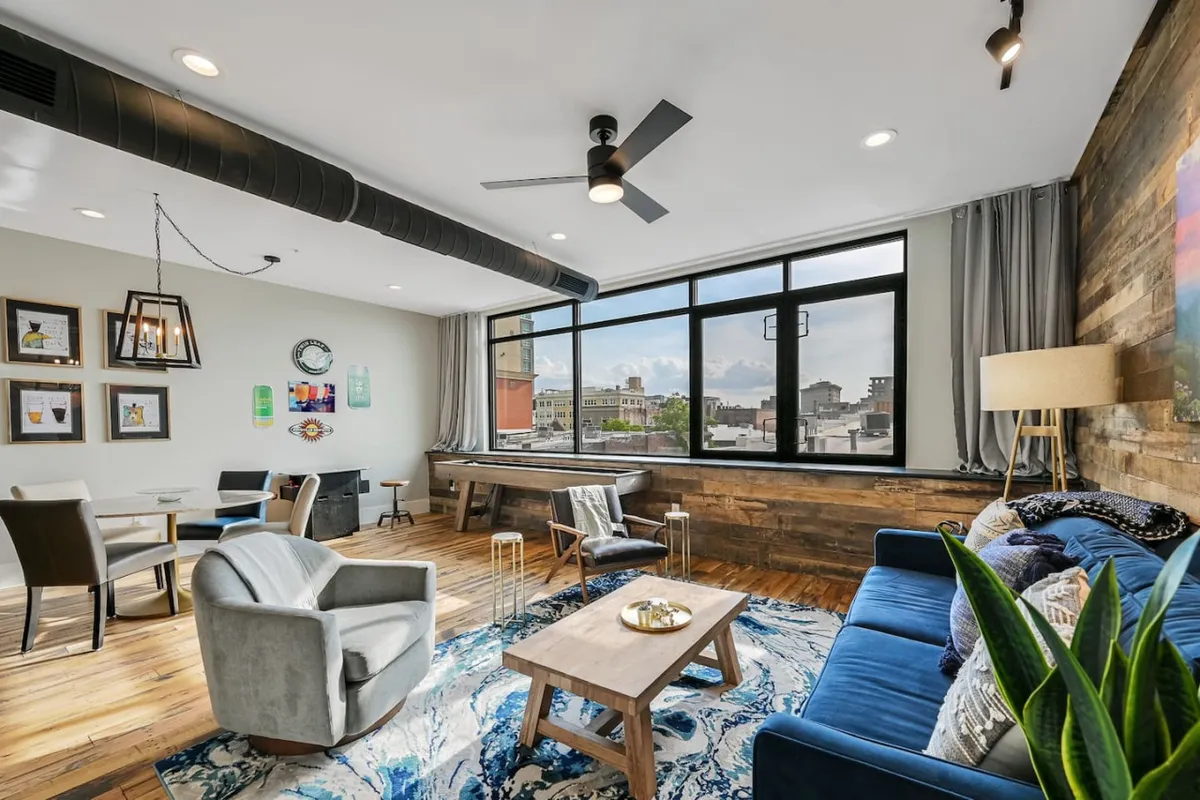 A stylish, sunlit living room on the third floor featuring expansive windows with sweeping downtown Asheville views at sunset. The space blends contemporary and classic design elements, with a plush blue sofa, chic wooden coffee table, and curated artwork creating an inviting atmosphere.