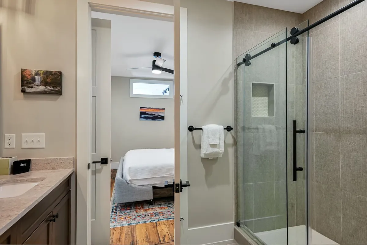 A modern bathroom featuring a glass shower enclosure, sleek vanity, decorative wall art, and a subtle view of the adjoining bedroom. Natural light enhances the refreshing and tranquil mood, ideal for peaceful mornings or relaxing evenings.