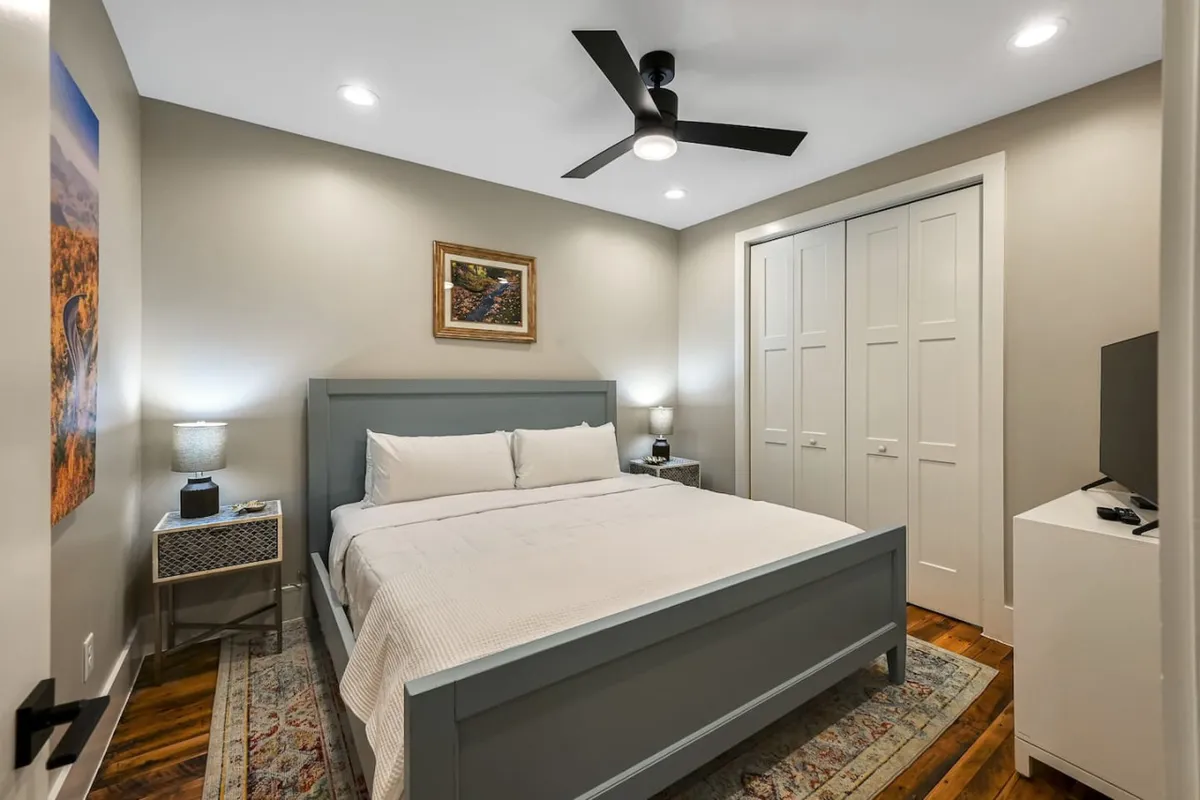This image features a beautifully made king-size bed framed by elegant nightstands, modern artwork above the headboard, and a closet in the background. The room is bathed in soft morning light, creating a calming and restful atmosphere. The decor is modern with subtle artistic touches that add personality without overwhelming the space.