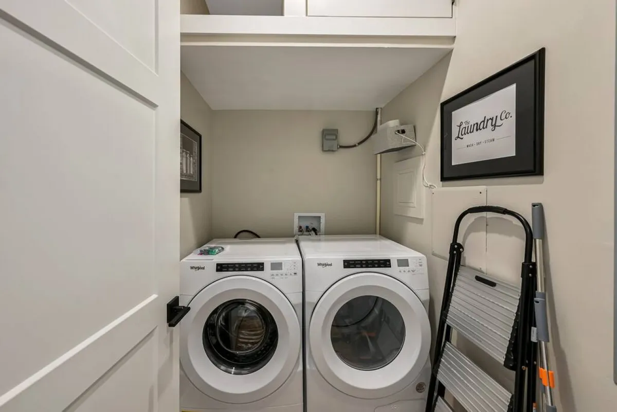 The image features a well-organized laundry area with a washing machine, dryer, a decorative 'laundry' sign, and a ladder. The space is clean, functional, and signals a sense of order and security, enhanced by the tidy arrangement of household items.